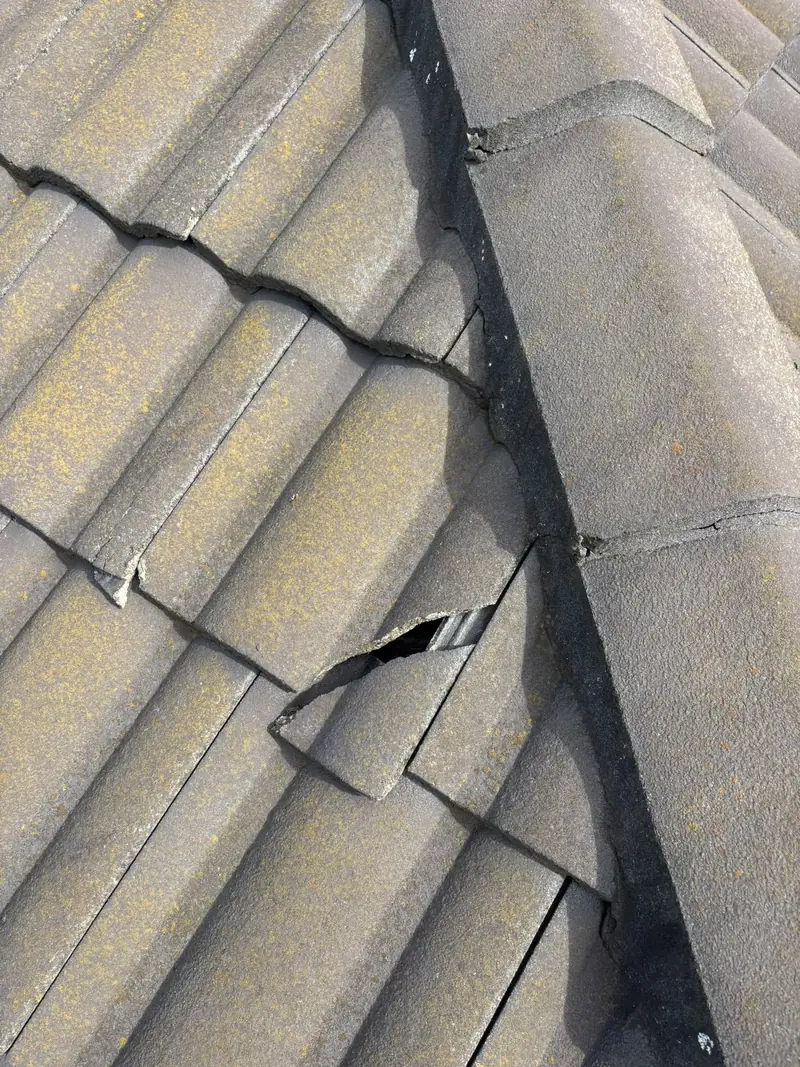 Cracked roof tile with a hole beside the ridge capping, lichen growing on the surrounding tiles